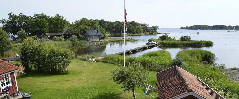 Röda stugor och en gräsmatta med flaggstång ner mot en vik med vass, en brygga och skogklädda öar