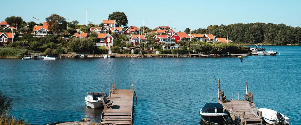Två träbryggor med små motorbåtar vid en klippig strand, och röda och vita hus bland träd på andra sidan en blå vik