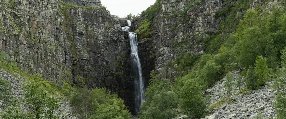 Smalt vattenfall som störtar ner i en djup klyfta mellan branta klippväggar, med grön björkskog och stenblock nedanför