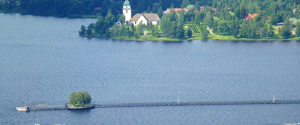 Lång träbrygga ut i en blank sjö, med en vit kyrka på en grön udde och skogsklädda stränder i bakgrunden