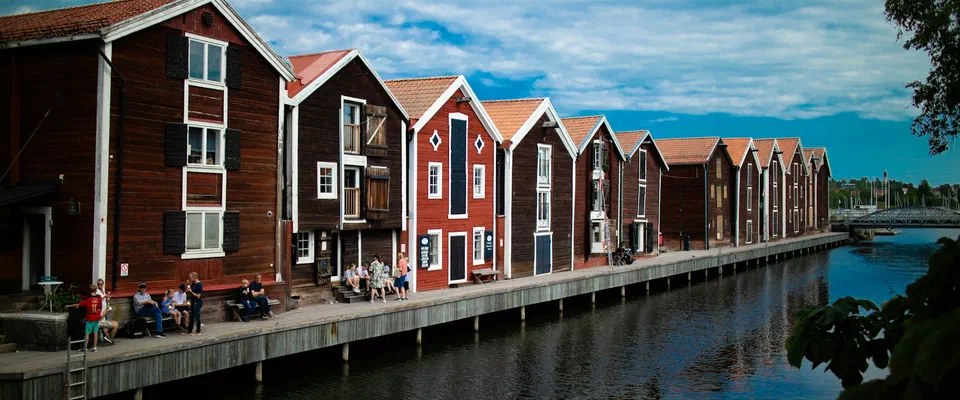 En lång rad röda och bruna sjöbodar i trä med vita knutar längs en kaj, som speglar sig i vattnet under blå himmel med moln