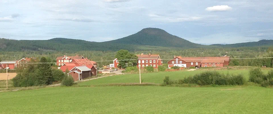 Röda gårdar på en grön åker i en dalgång, med ett kupolformat skogsberg i bakgrunden under blå himmel med vita moln