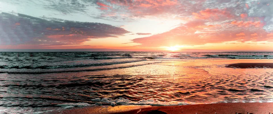 Solen går ner över havet bakom låga vågor på en sandstrand, under en himmel med rosa och gråblå moln
