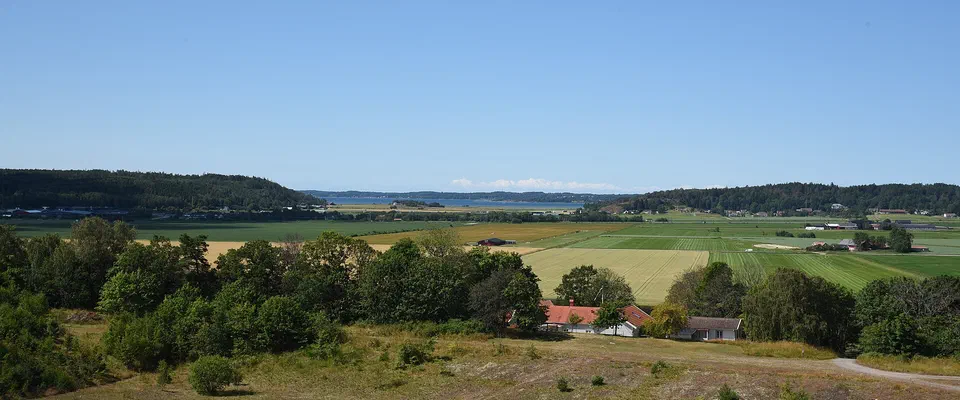 Utsikt från en höjd över buskmark, en liten damm och ett rött hus mot åkrar, skog och en fjord i fjärran under blå himmel