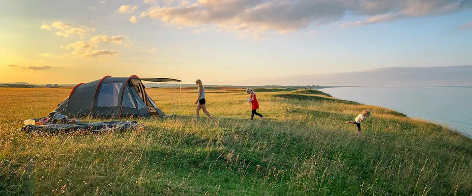 Grått tunneltält på en gräsbevuxen strandbrink vid havet i kvällssol, med barn som springer i gräset