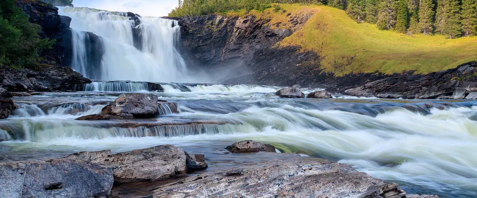 Brett vattenfall som forsar över mörka klipphällar, med granskog och en solbelyst gräsbrink bredvid