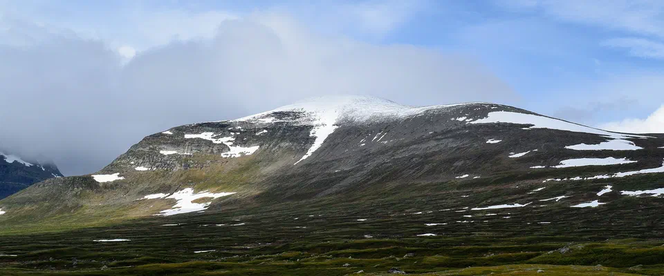 Brett fjäll med snöfläckar på toppen och molntäcke, ovanför en grön fjällhed