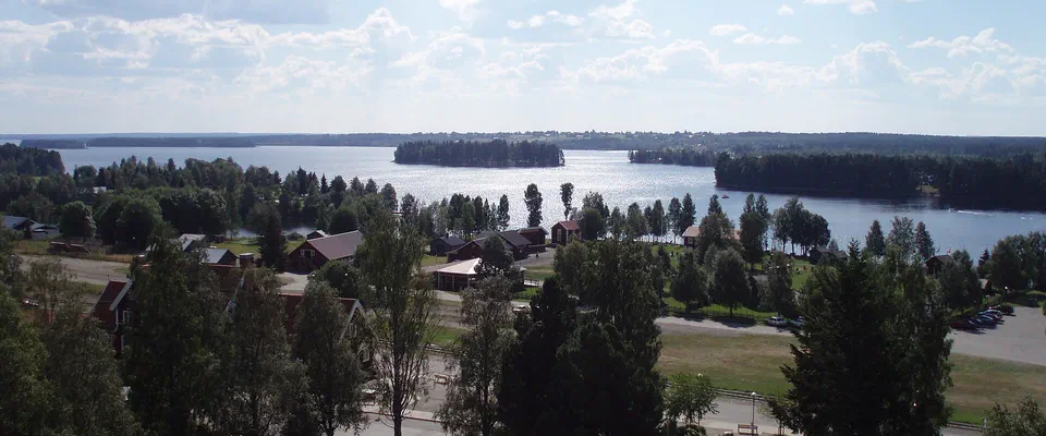 Utsikt över röda trähus och lövträd ner mot en stor sjö med skogsklädda öar under en sommarhimmel med vita moln
