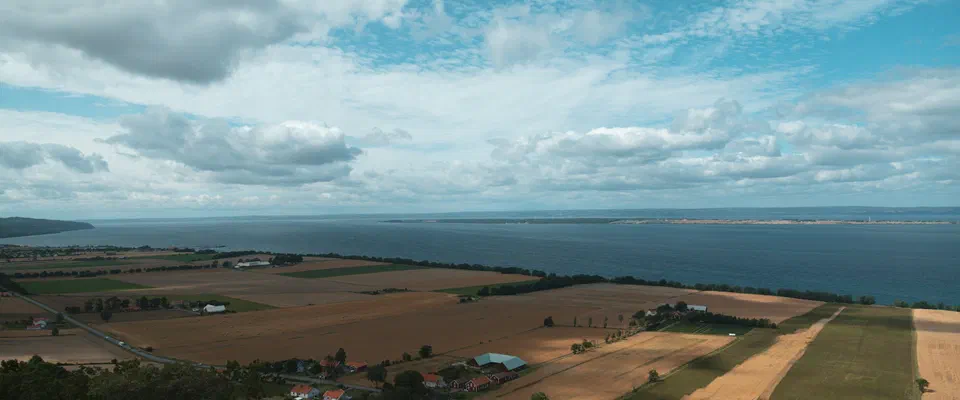 Utsikt från en höjd över gröna och gula åkrar och gårdar ner mot den stora sjön Vättern under en blå himmel med moln