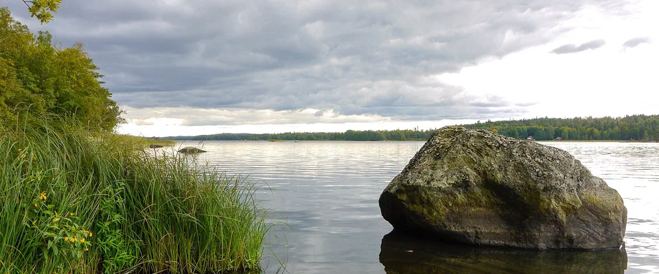 Ett stort mossbevuxet stenblock som speglar sig i en blank sjö, med vass och gräs i förgrunden och skog på andra stranden