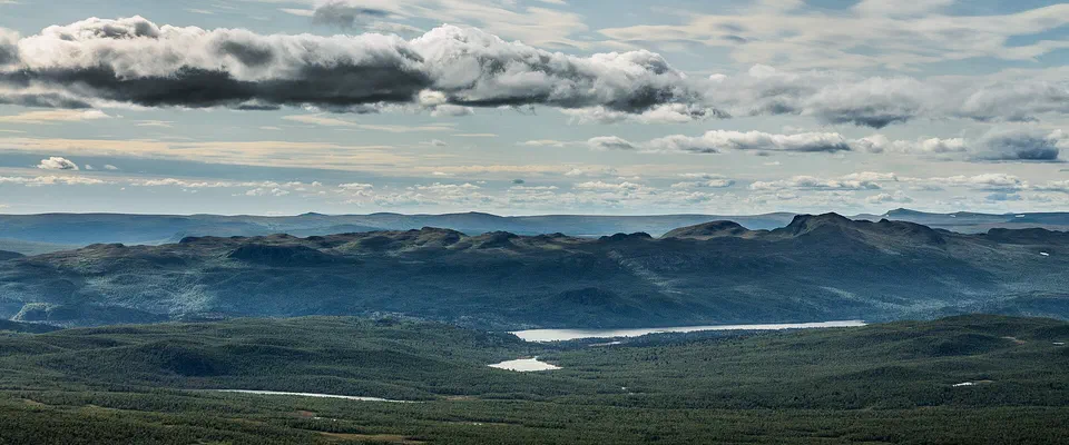 Vid utsikt från ett fjäll över skogklädda dalar med en avlång sjö och en rad blågrå fjäll bakom, under en himmel med långa moln