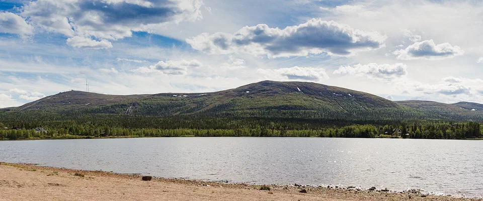 Fjället Dundret med snöfläckar på toppen, sett över en blank sjö med sandstrand i förgrunden