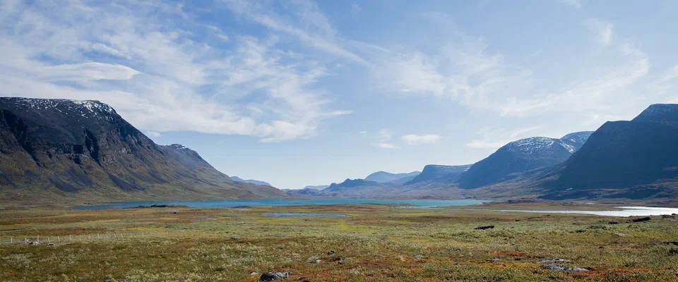 Grön och rödbrun fjällhed som sluttar ner mot en sjö, med branta fjäll på båda sidor under blå himmel med tunna moln