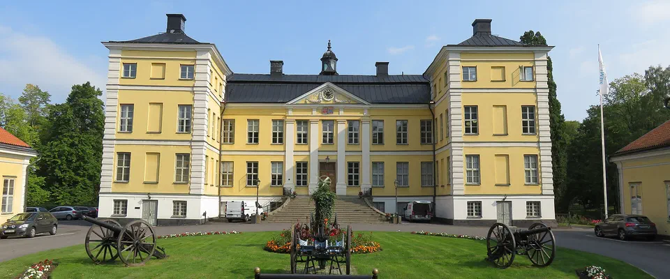 Gult slott i tre våningar med svart tak och flyglar, med kanoner och en rund gräsplan framför
