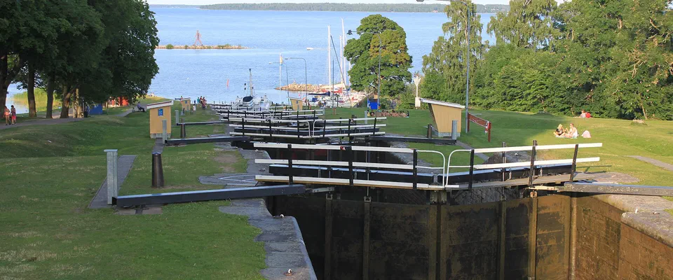 Slusstrappa med svarta slussportar och grönt gräs, ner mot en sjö med båtar och lövträd
