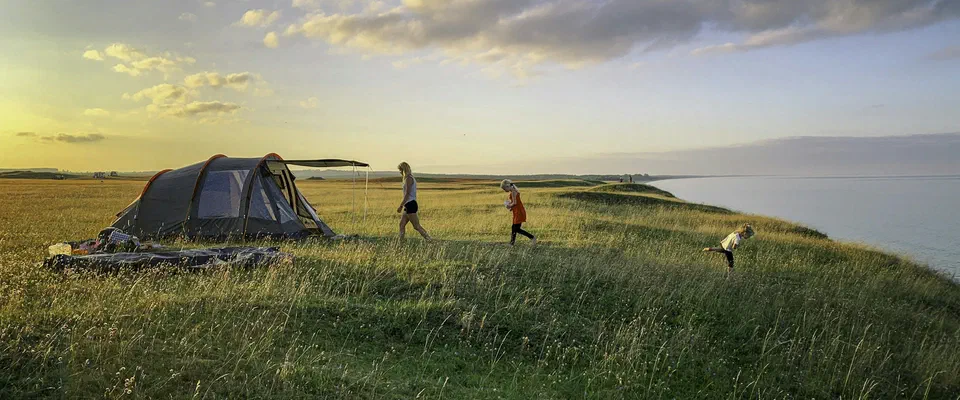 Ett tält på en grön äng ovanför havet i kvällssol, med barn som springer i gräset under en himmel med moln