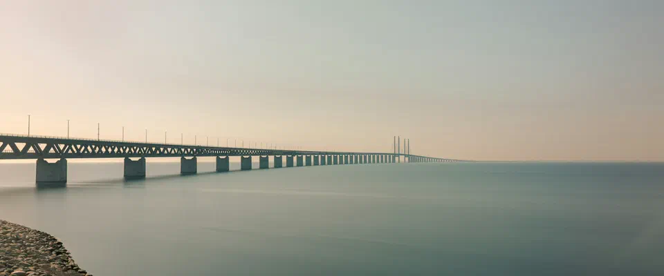 Den långa Öresundsbron över ett stilla, grönblått sund, sedd från en stenig strand i disigt kvällsljus