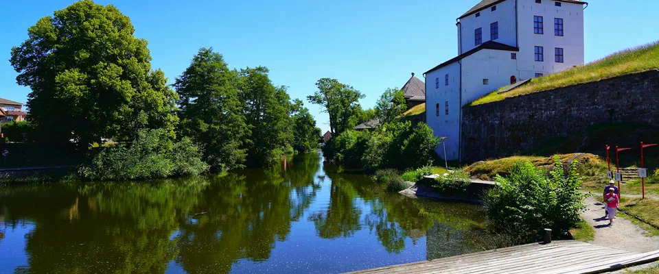 Vitt slottstorn och gamla murar bakom lövträd vid en å som speglar den blå himlen, med en gångväg längs vattnet