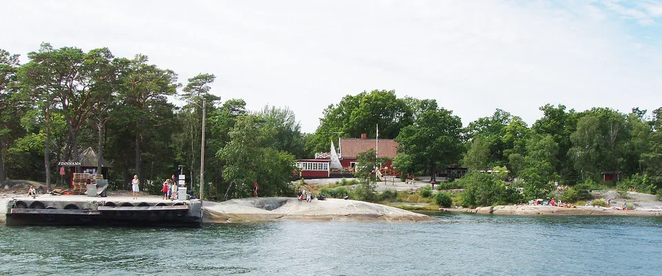 Brygga och strand med café och röda hus bland lövträd på en skärgårdsö en ljus sommardag