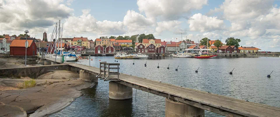 Träbrygga på betongpelare över släta klippor ut i en vik, med en småstad av röda och gula hus och båtar i bakgrunden