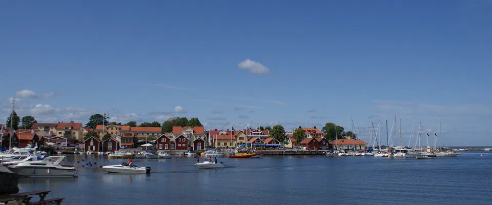 Vik med motorbåtar och segelbåtar framför röda och gula trähus under en klarblå himmel, med en picknickbänk på klipporna i förgrunden