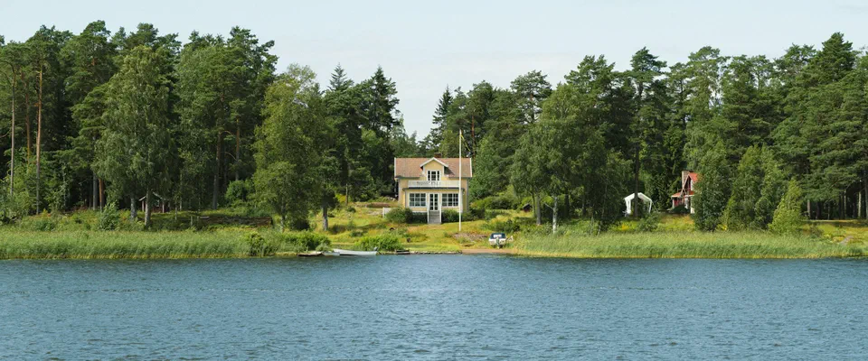 Blått vatten framför en strand med lövskog och ett vitt hus bland träden, under klarblå himmel