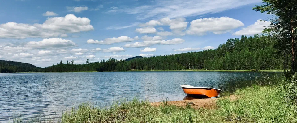 Orange roddbåt uppdragen på en gräsbevuxen sandstrand vid en sjö med skogklädda stränder, under blå himmel med vita moln