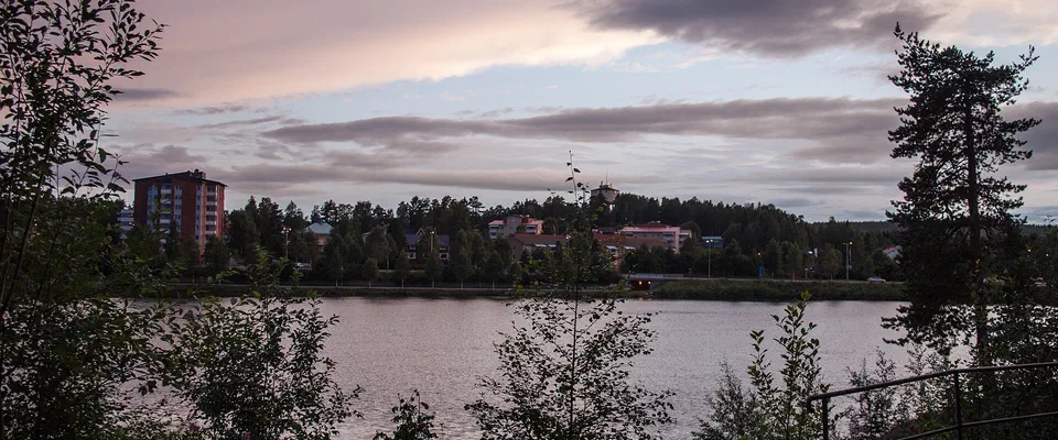 Lugn älv i rosa kvällsljus med björkar i förgrunden och hus på andra stranden