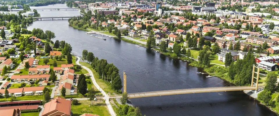 Flygbild över en bred älv med en gångbro, villor och flerbostadshus på båda stränderna och staden bakom
