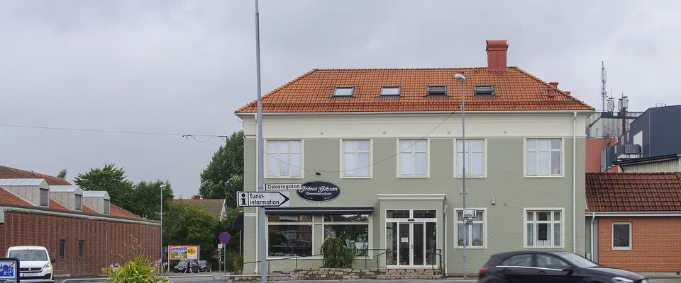 Ljusgrönt tvåvåningshus med tegeltak i en gatukorsning, med blomsterurna och parkerade bilar
