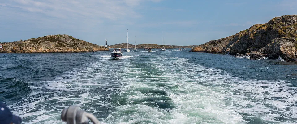 Skumsvall efter en båt mellan släta granitklippor och skär, med segelbåtar och en fyr i fjärran