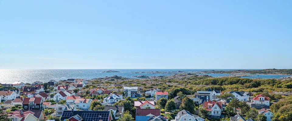 Flygbild över ett tätt samhälle med vita och röda hus bland klippor och grönska, med havet bakom