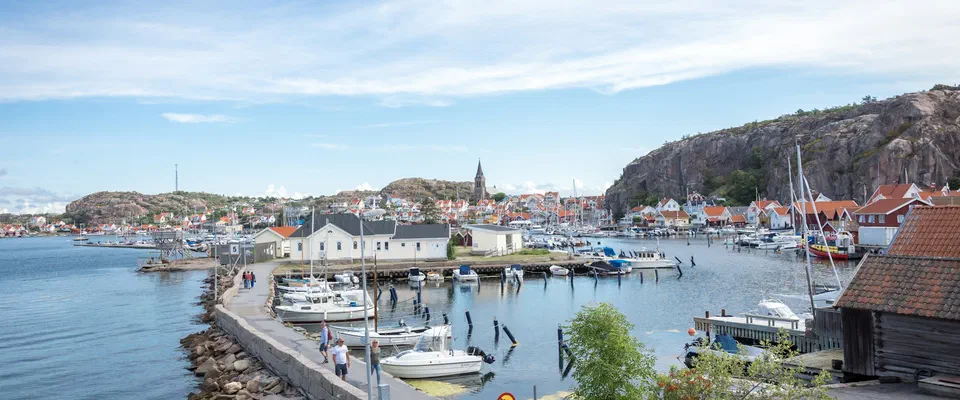 Hamn full av motorbåtar och segelbåtar längs en stenpir, med vita trähus och en brant klippa till höger under blå himmel