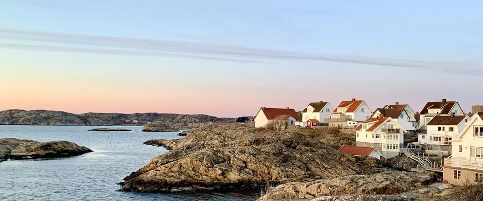 Vita och röda trähus på kala granitklippor vid en lugn havsvik i kvällsljus, med små skär i vattnet