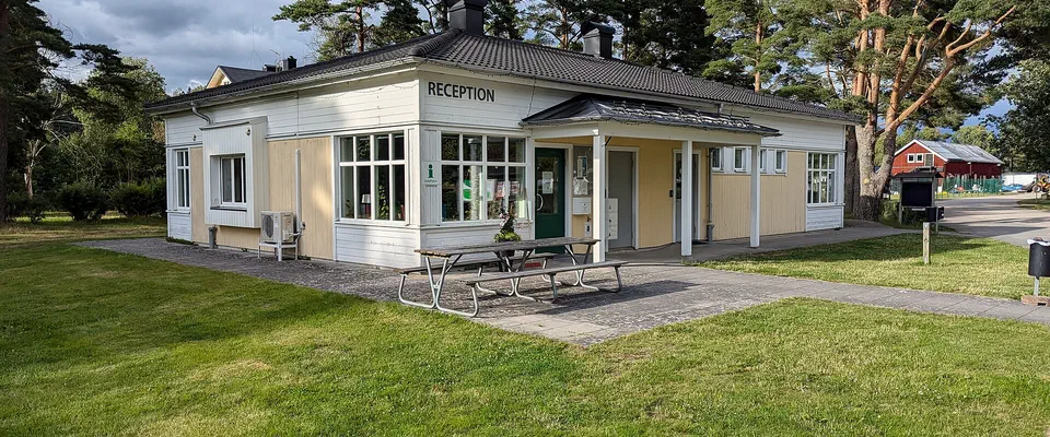 Ljusgul envåningsbyggnad med skylten Reception vid en gräsmatta och höga tallar
